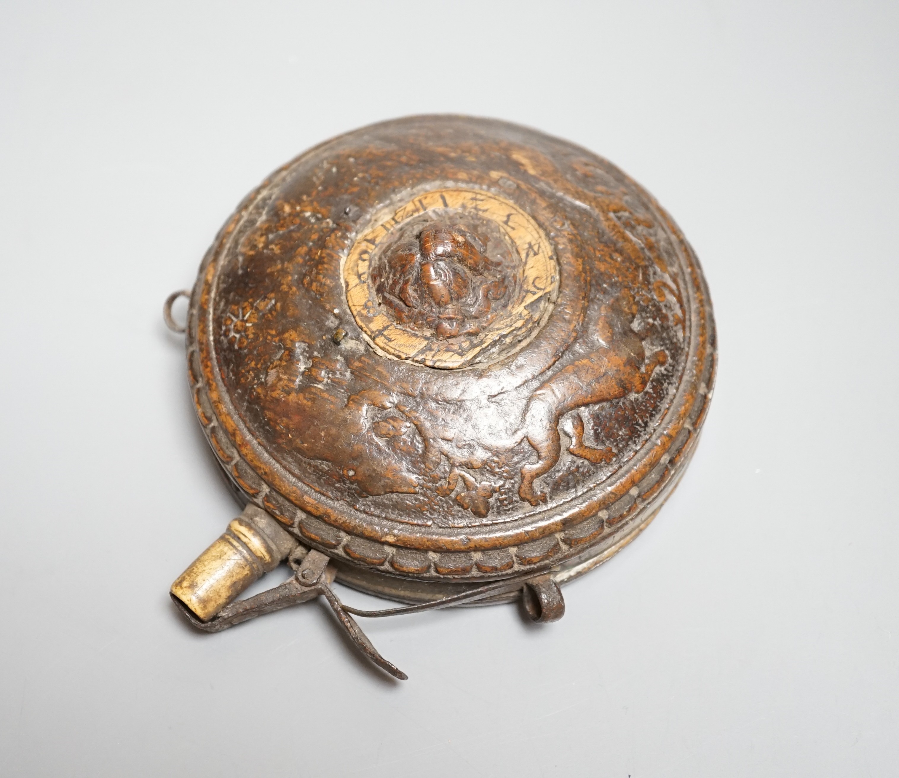 A circular wooden powder flask 17th century, carved with bear, hound and fox, and lion’s mask in low relief surrounded by game counter, engraved brass border, iron lever charger, turned bone nozzle. Diameter 11cms